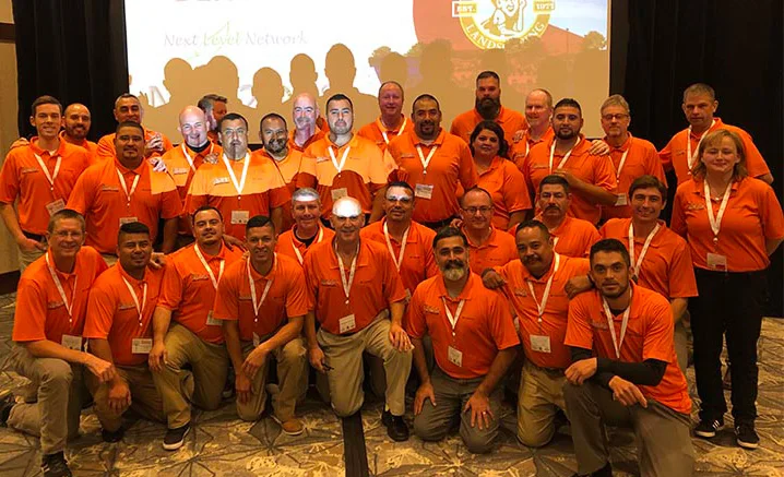 A group of 34 people, wearing orange shirts with lanyards, posing for a photo in what appears to be a conference or meeting room. A screen with a logo is in the background.