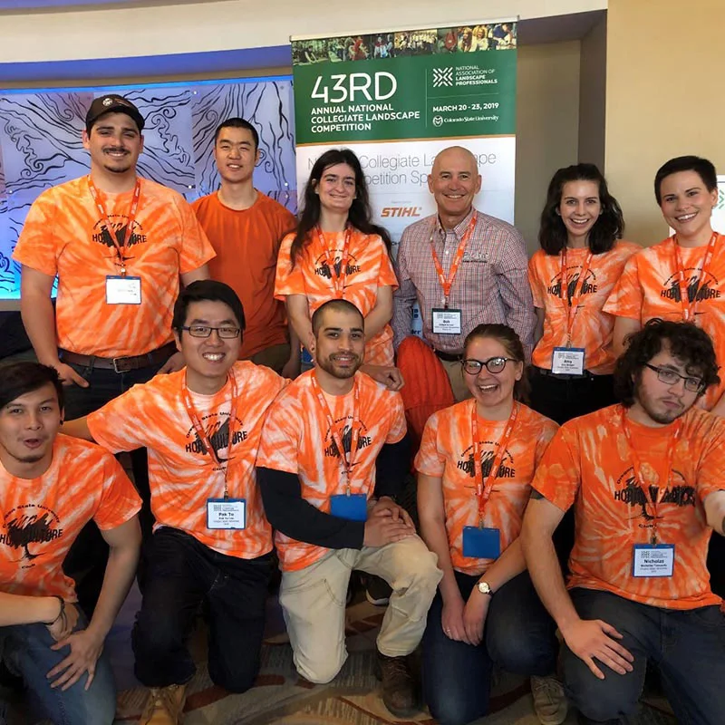 A group of 11 people wearing orange tie-dye shirts and name tags pose in front of a banner for the 43rd Annual National Collegiate Landscape Competition. The event occurred March 20-23, 2019.