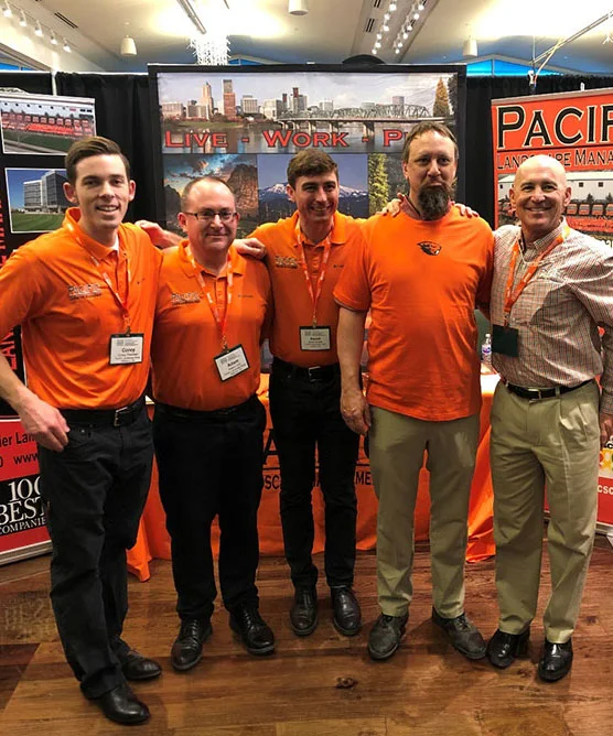 Five men, four in orange shirts and one in a checked shirt, stand smiling at a trade show booth with banners in the background. They each wear name badges and conference lanyards.