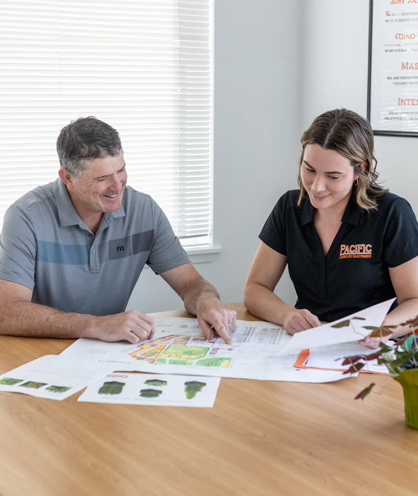 A man and a woman sit at a table reviewing a landscaping plan. The man points to a section on the plan while both of them smile. The woman wears a