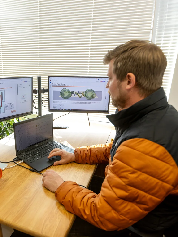 A man sits at a desk with a laptop and two monitors, reviewing architectural plans for Home Plate Center on his screens. He is in an office with window blinds partially open.