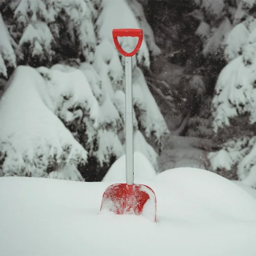Red snow shovel with metal handle partially buried in deep snow, standing vertically in a snowy landscape with snow-covered trees in the background.