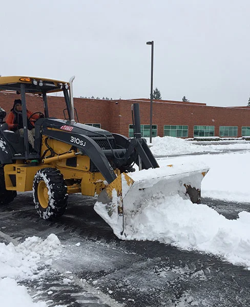 A yellow and black construction vehicle is clearing snow from a parking lot in front of a brick building with green windows. The sky is cloudy, and the ground is covered with snow.