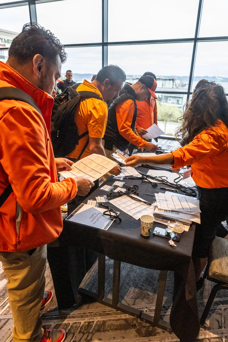 People in orange jackets and shirts are gathered at a registration table, checking documents and name tags. The setting appears to be indoors with a large window in the background.