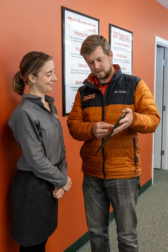 Two people stand in a hallway, with three framed motivational posters behind them. The man in an orange jacket shows the woman in a gray shirt something on a tablet.