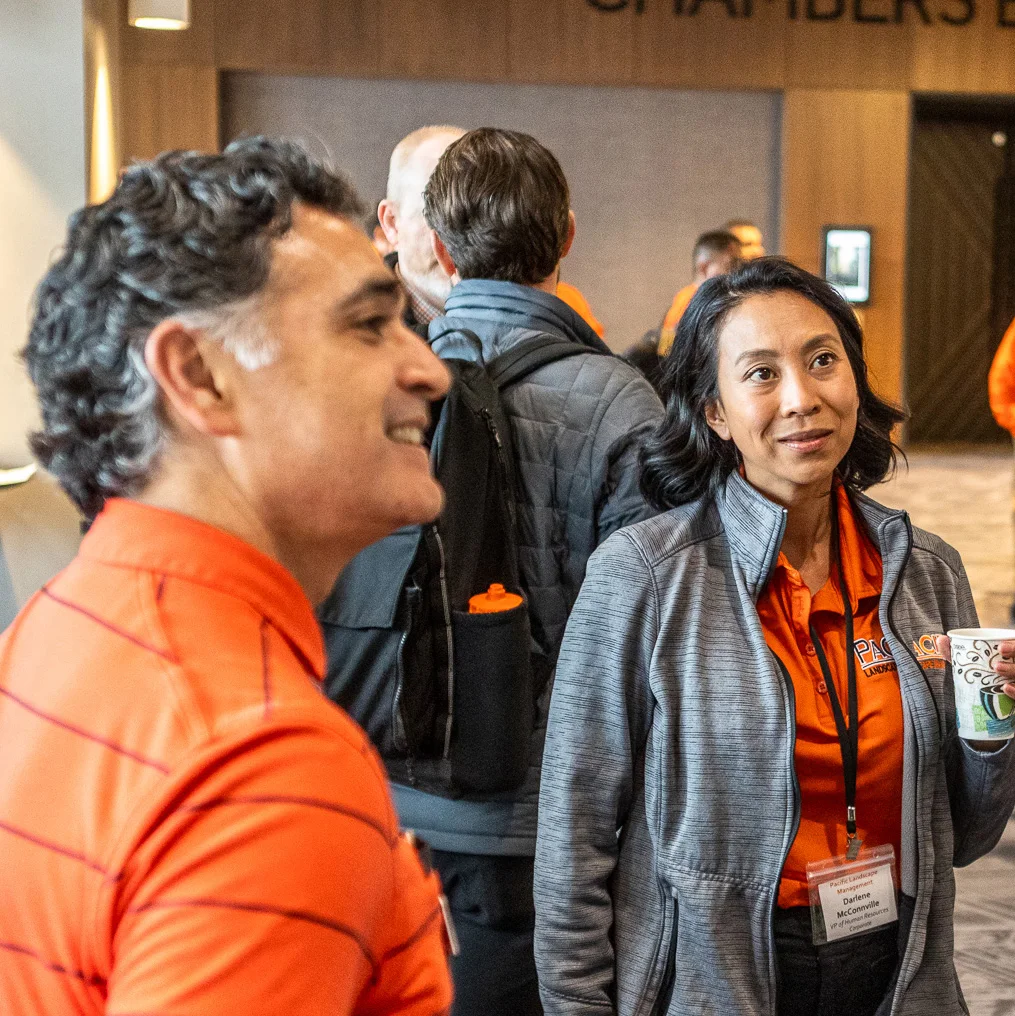 Two people in orange shirts with name tags converse at a networking event inside a conference room. There's a group of people in the background, one person holding a cup in the foreground.