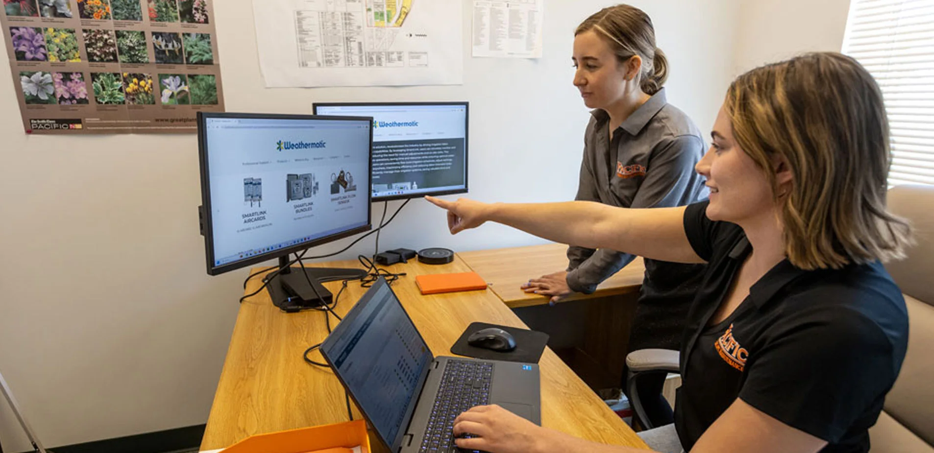 Two women working at a desk, looking at two computer monitors displaying the Weathermatic website. One woman points at the screen while the other works on a laptop.