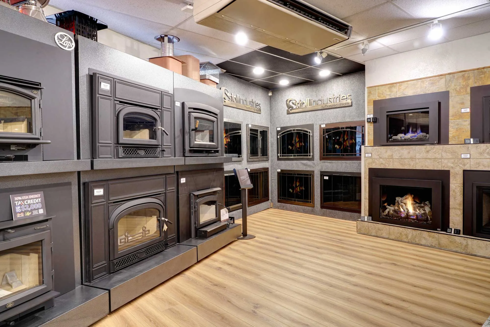 Showroom with various fireplace models on display. Includes traditional and modern designs, with black and metallic frames, positioned against gray and beige walls. Wood flooring beneath.