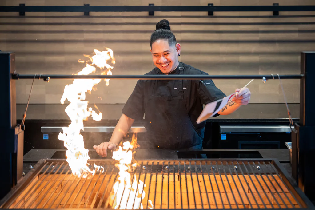 Chef smiles while grilling food over an open flame in a kitchen, holding a utensil in one hand. The kitchen is modern, with clean lines and sleek surfaces in the background.