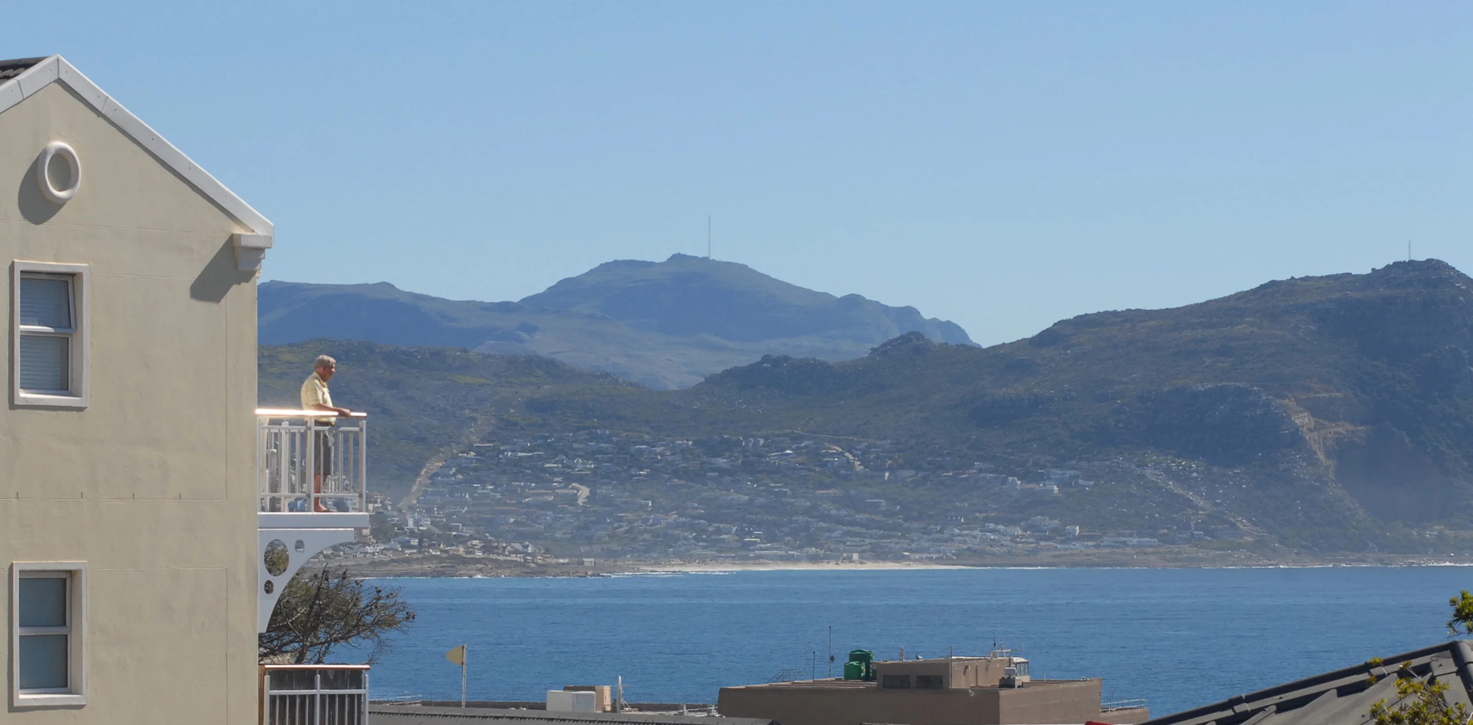 A person stands on a balcony overlooking a body of water with mountains in the background. The building is cream-colored with two windows visible. The sky is clear and blue.