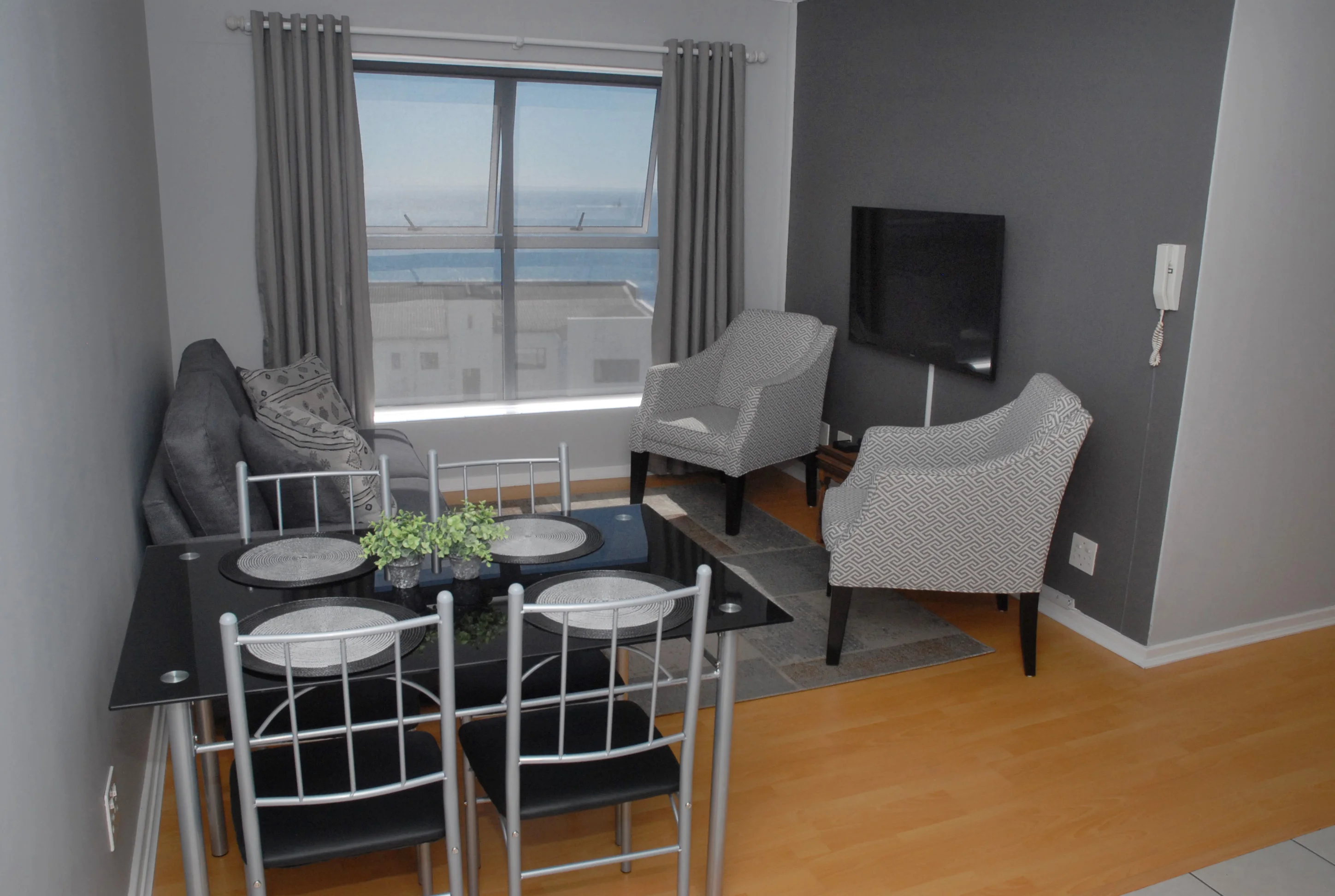Modern living room with a glass dining table set for four, two patterned armchairs, a sofa, and a wall-mounted TV. A large window shows a view of the ocean.