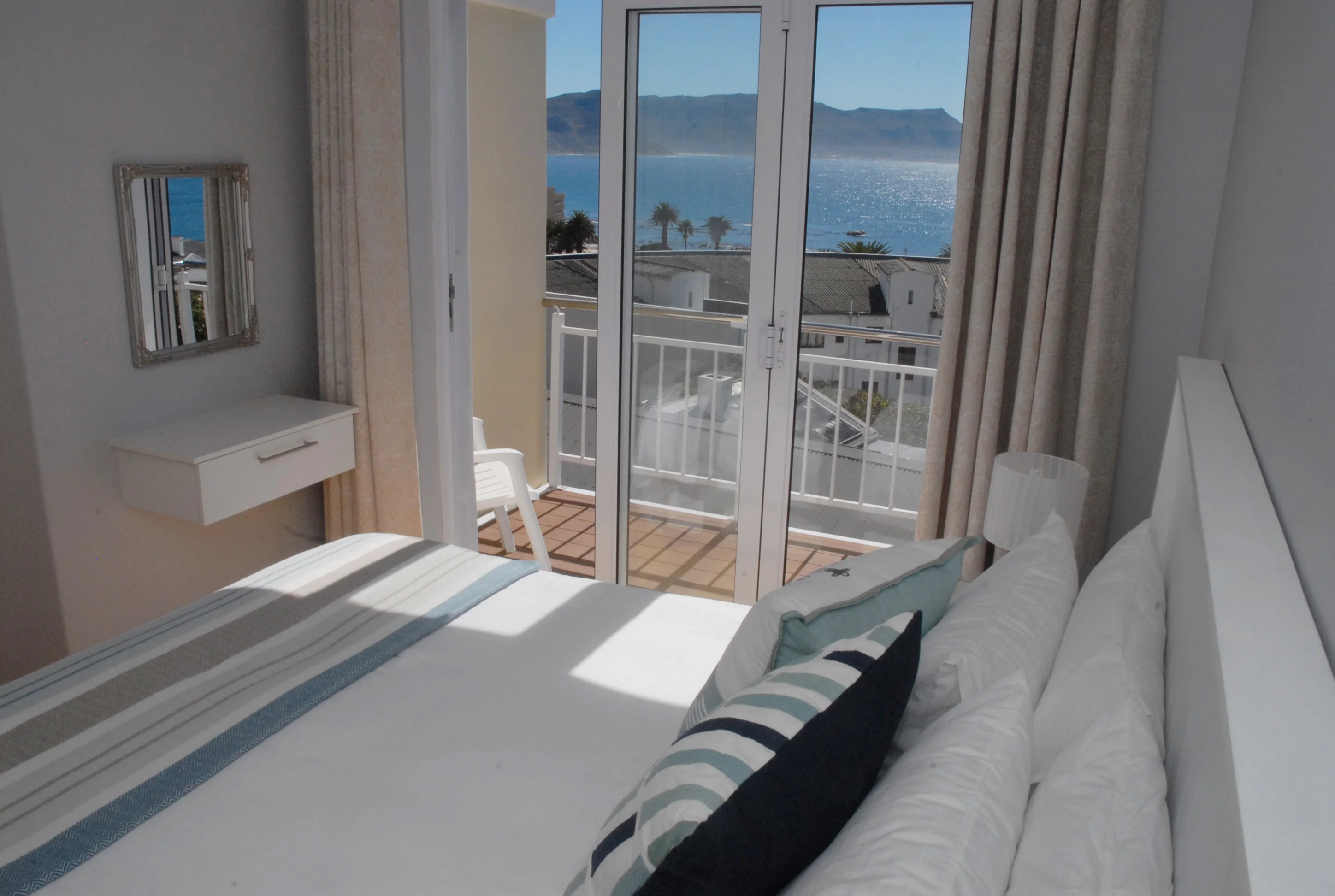 Bright bedroom with a bed, striped pillows, and a small wall-mounted desk. Glass doors open to a balcony with ocean view, mountains in the distance, and a clear blue sky.