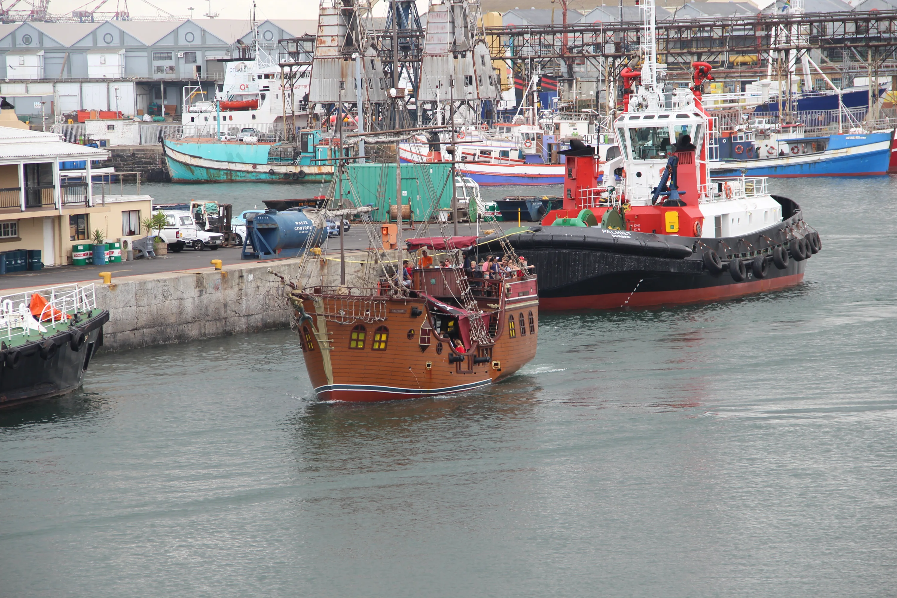 A wooden pirate ship with yellow windows sails in a busy harbor near a red and black tugboat. Industrial buildings and various boats are visible in the background.