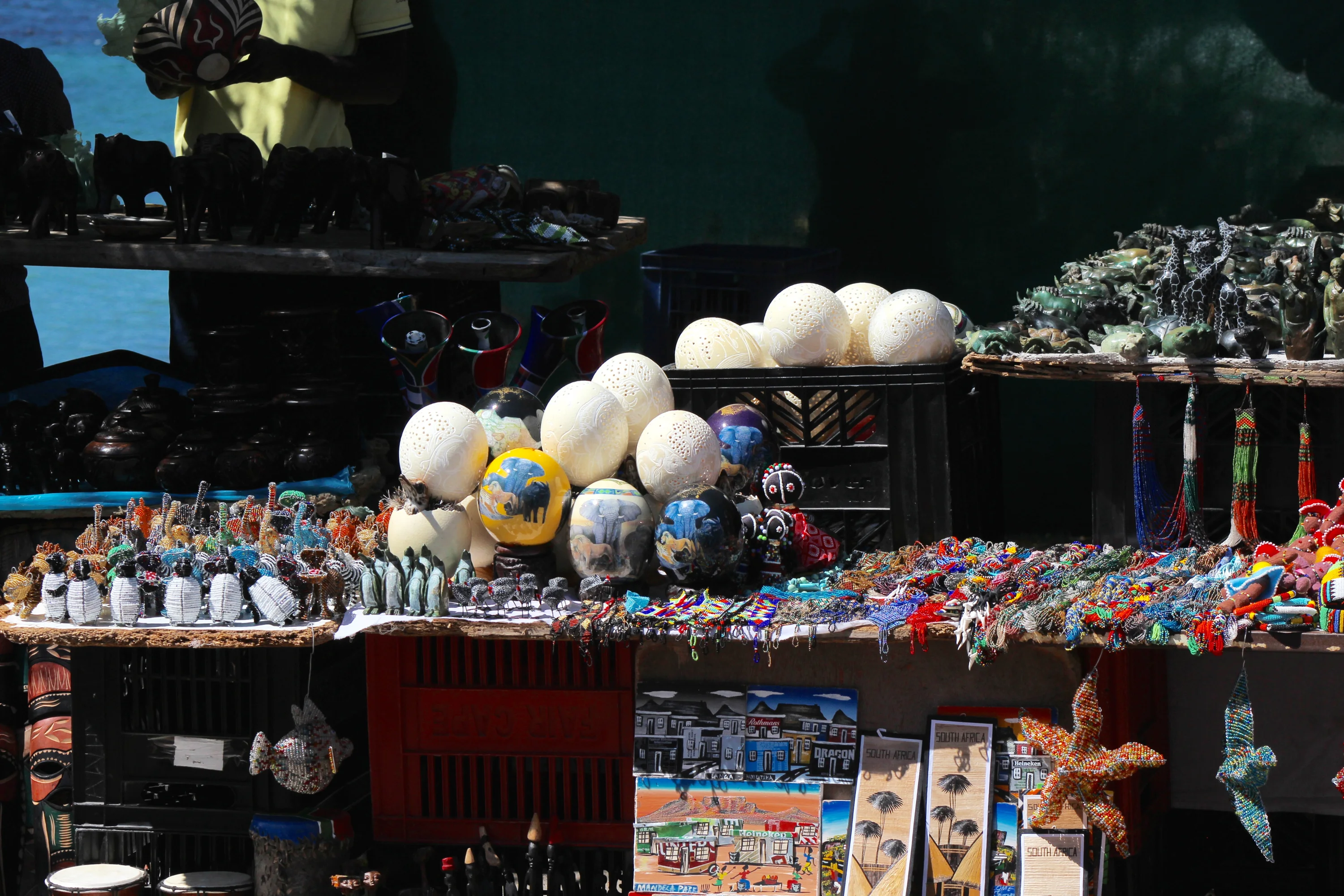 A market stall displays various handcrafted items, including beaded figurines, painted eggs, colorful textiles, and artwork under a shaded area with a lake in the background.