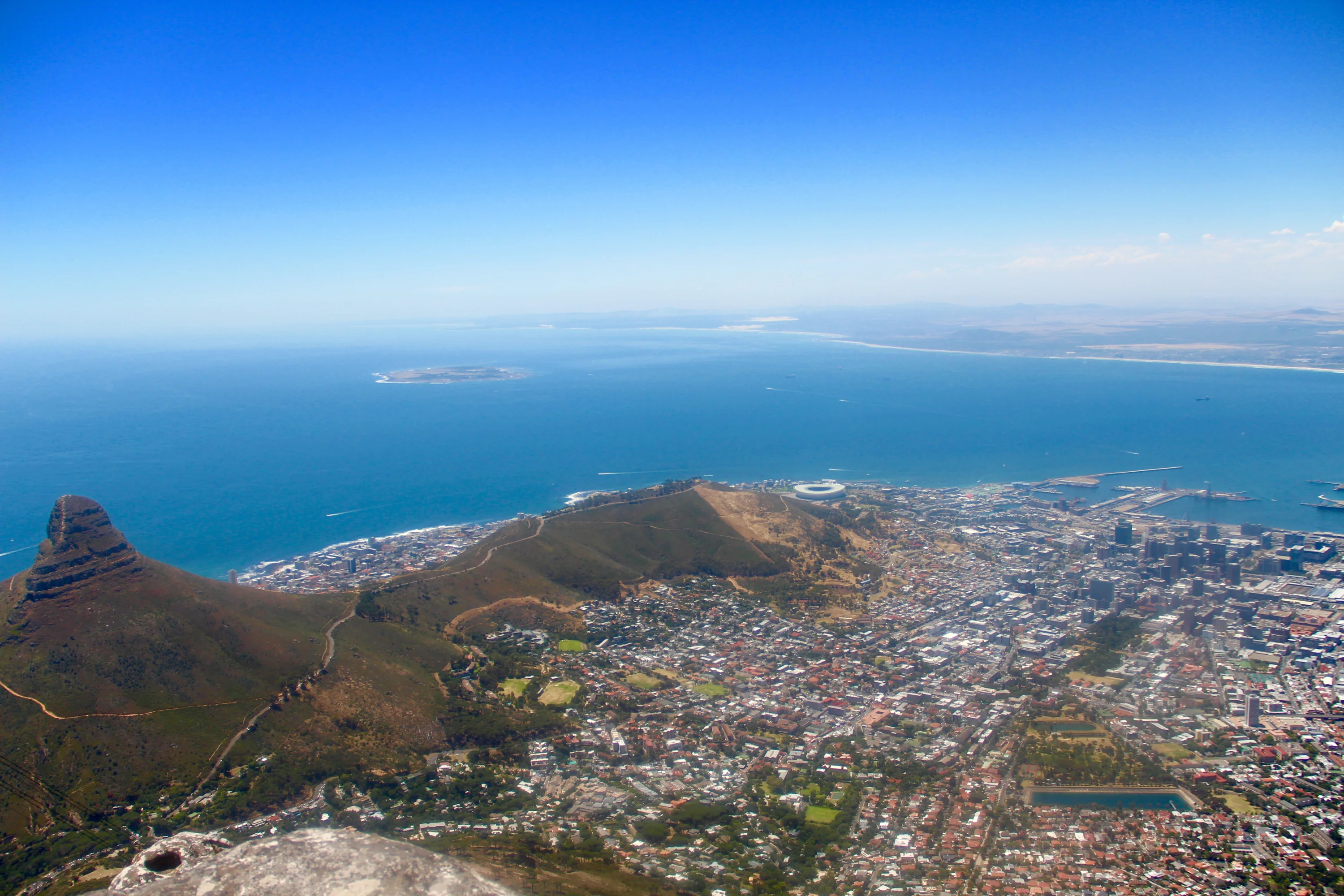 Aerial view of Cape Town, South Africa, featuring Lion's Head mountain, the cityscape, and the surrounding coastline with Robben Island visible in the distance under a clear blue sky.