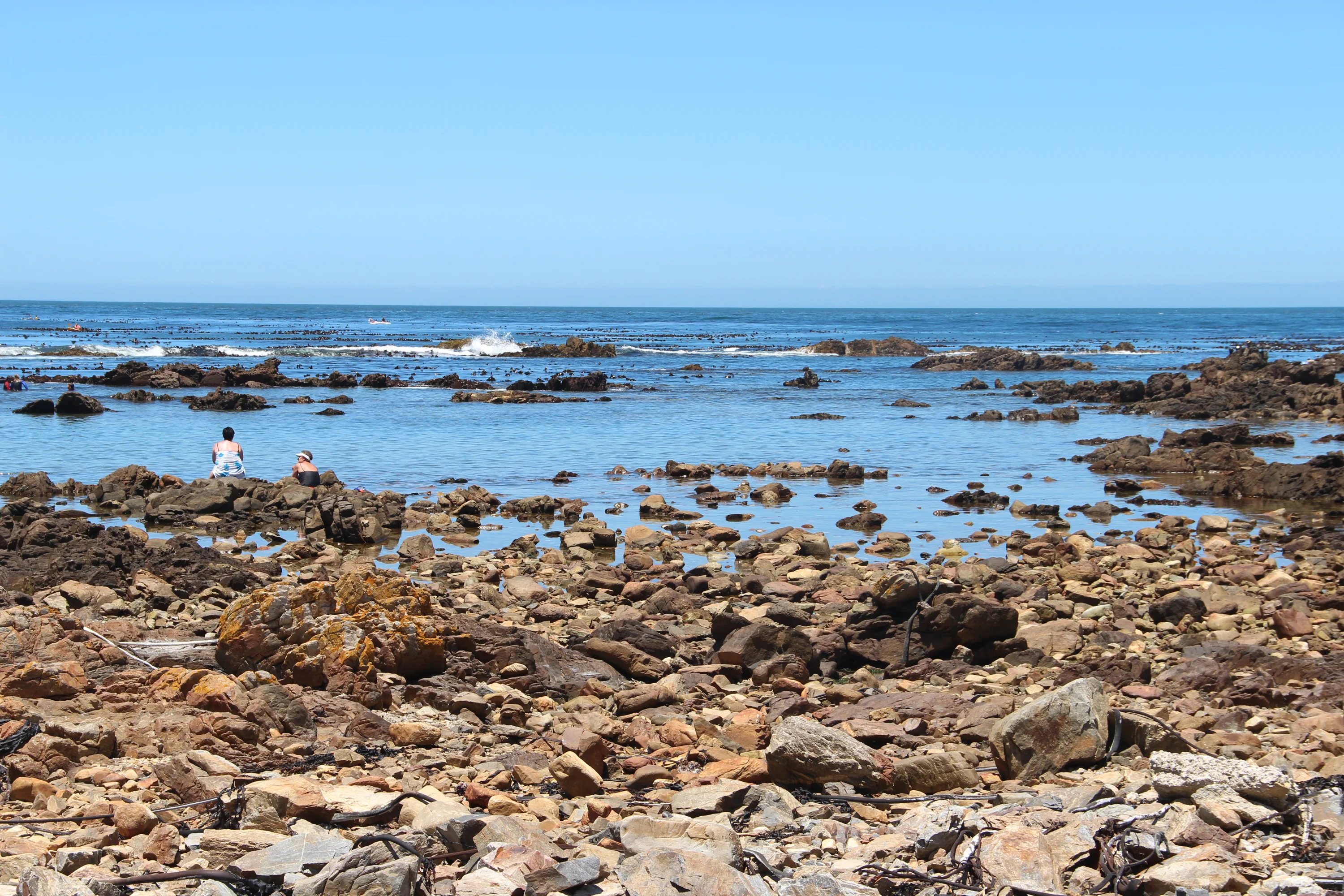 A rocky beach with two people sitting on rocks near the shoreline. The calm ocean extends to the horizon under a clear blue sky, with waves gently breaking on the rocks.