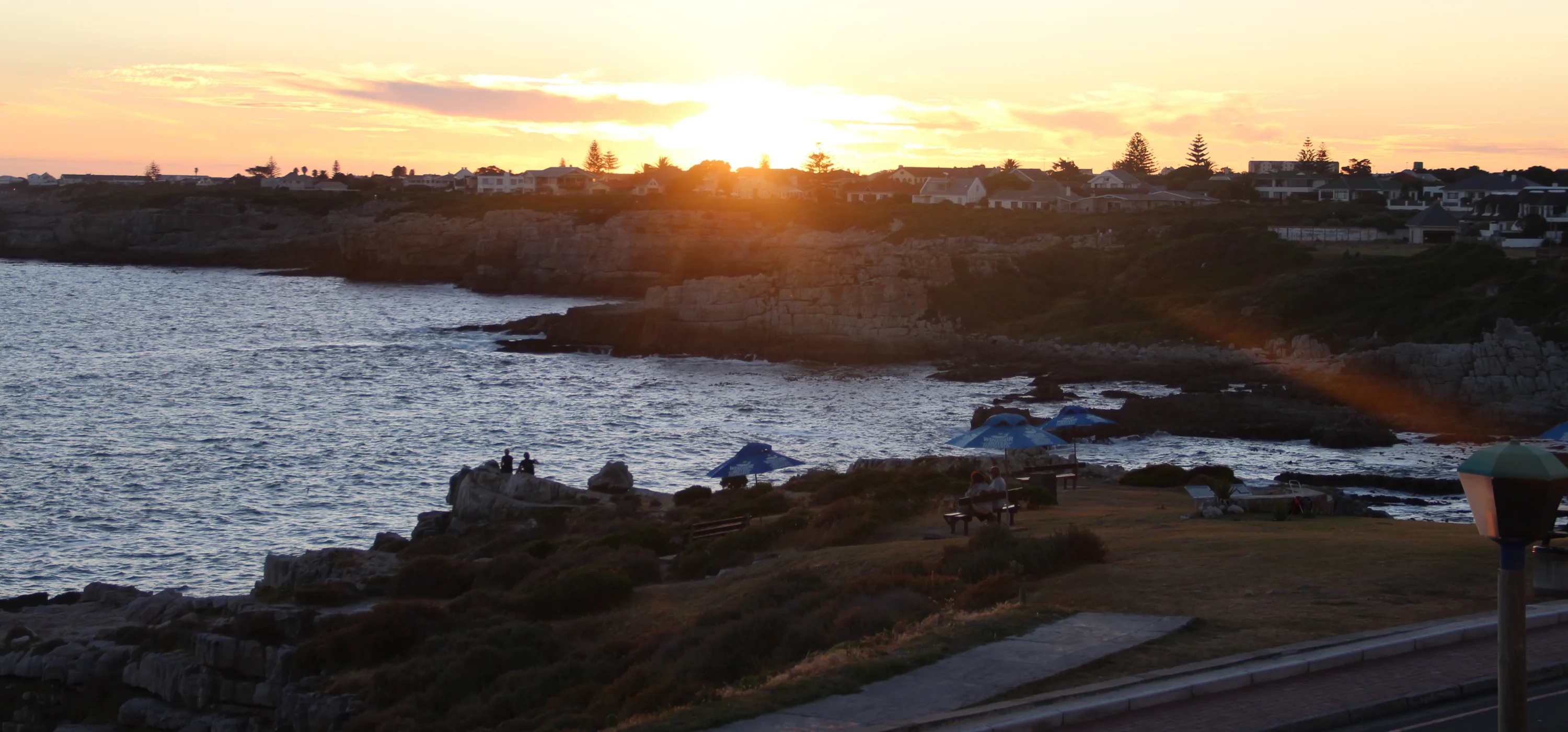 Sunset over a coastal town with rocky cliffs and ocean waves. Silhouetted figures sit on rocks, while others relax under umbrellas on grassy areas near a walkway.