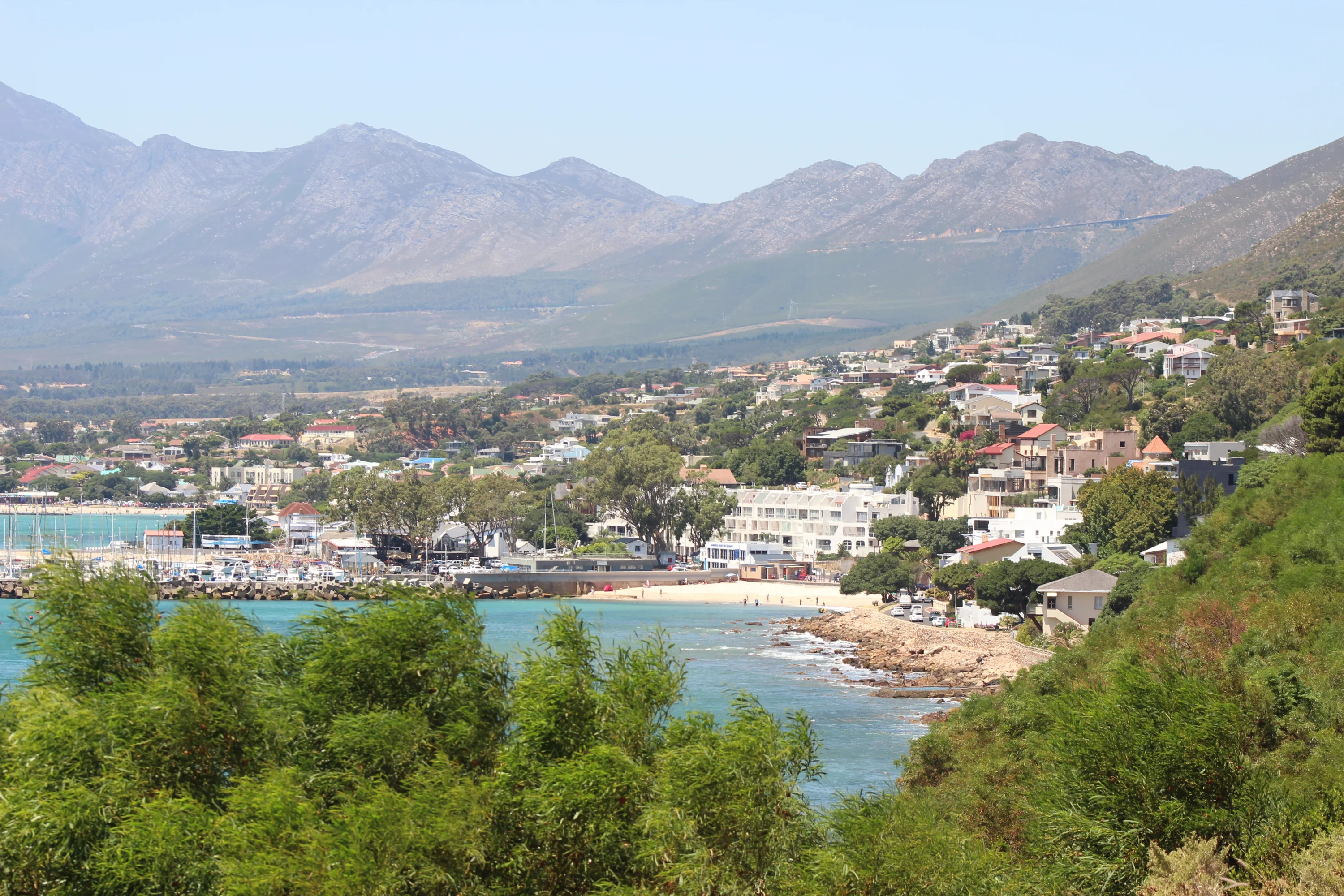 Coastal town with beachfront houses, surrounded by lush greenery and mountains in the background. The sea is calm with a small sandy beach and a marina visible.