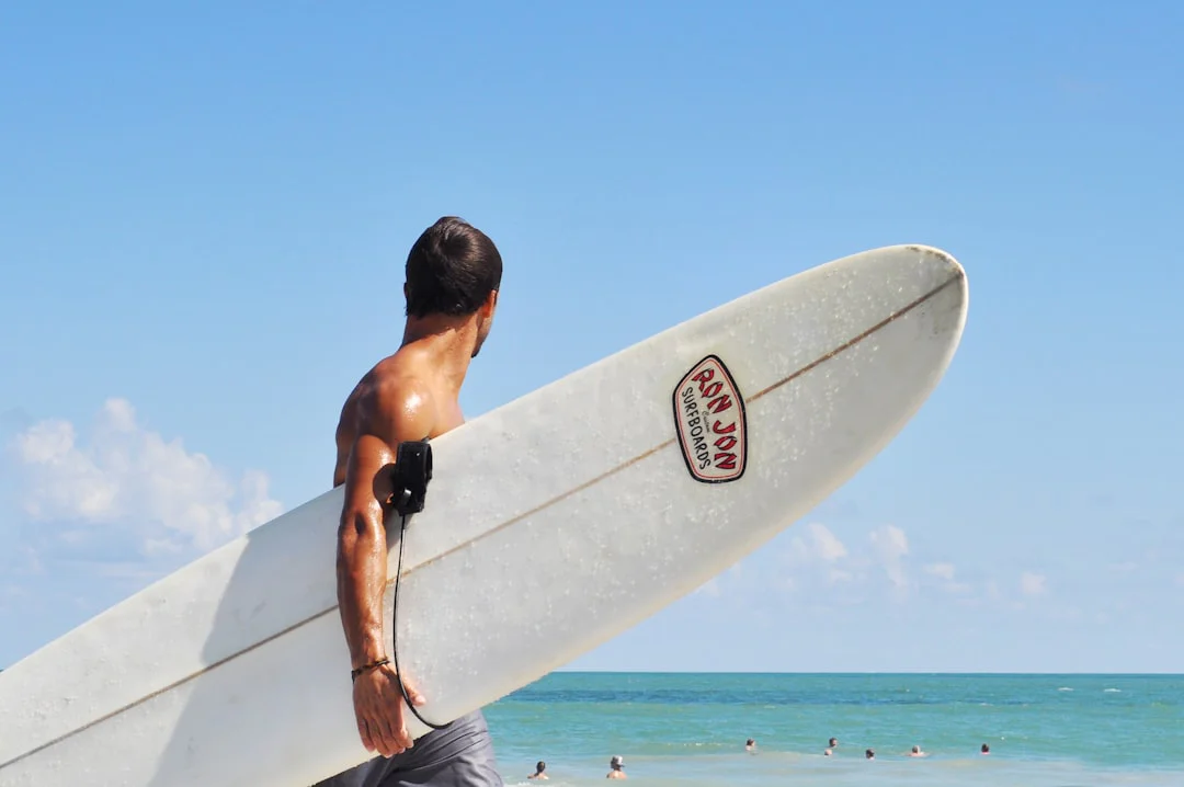 Surfer holding a Ron Jon surfboard, standing at the beach with a clear blue sky and ocean in the background. Several people are swimming in the water.