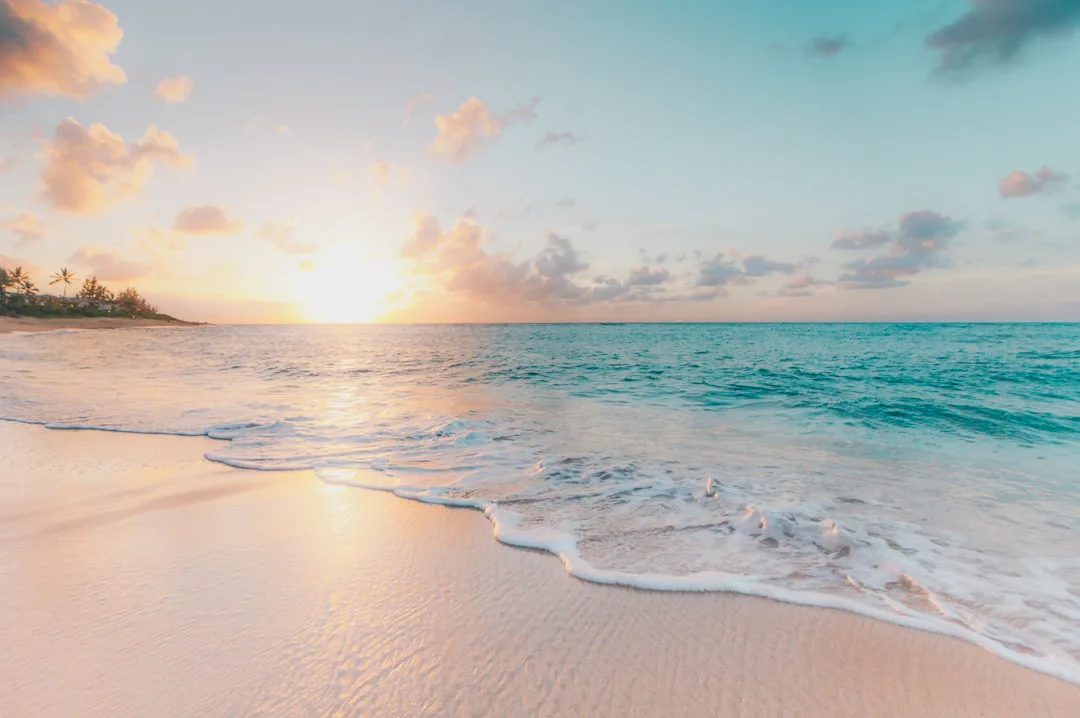 Sunset over a sandy beach with gentle waves lapping the shore, palm trees in the distance, and a sky filled with soft clouds tinged with pink and orange hues.