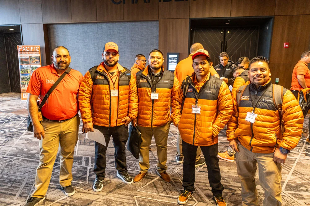 Five men wearing orange and black jackets, standing together and smiling at a conference. Additional people and a 