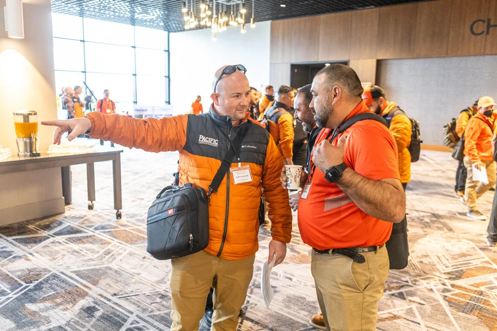A group of men in orange jackets converse inside a well-lit conference hall. One man is pointing while holding a document. Tables with refreshments are visible on the left.