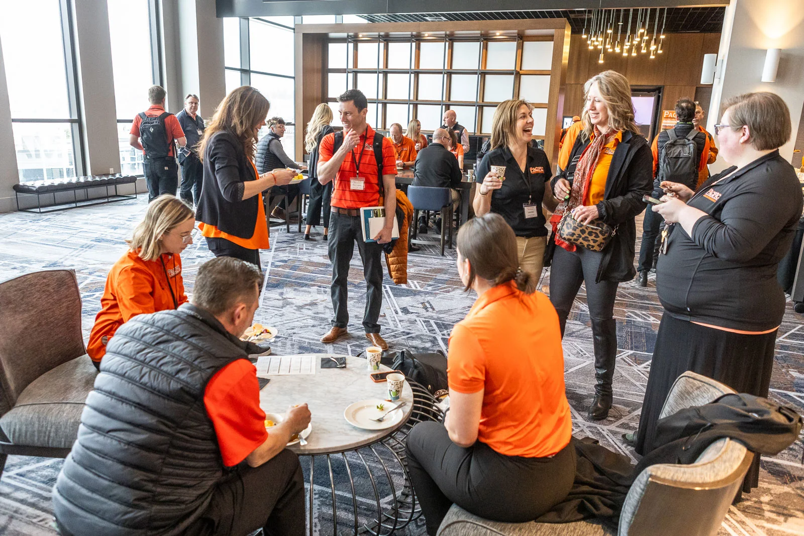 A group of people wearing orange and black shirts stand and sit around tables in a large, modern conference room, engaged in conversation and enjoying snacks.