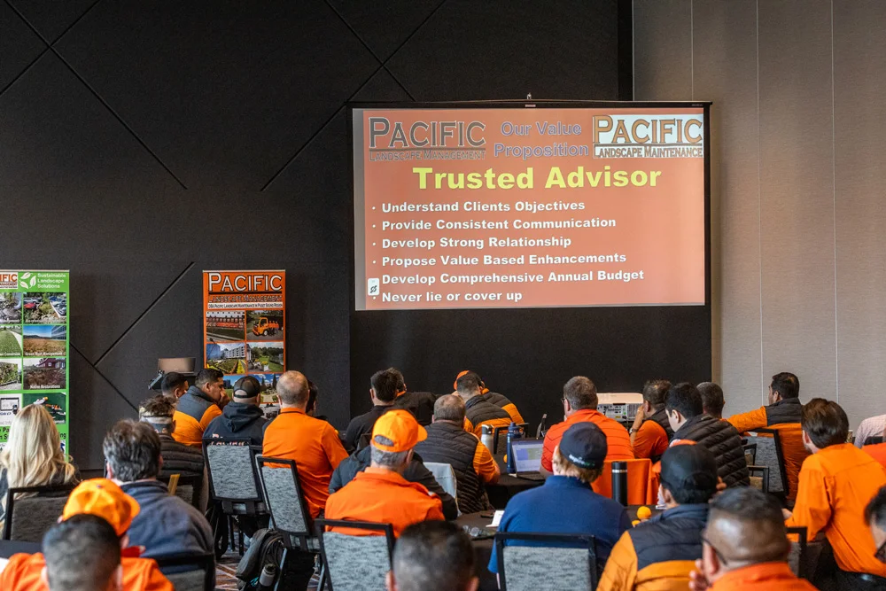 A group of people in orange shirts are seated and facing a large screen displaying a presentation titled 