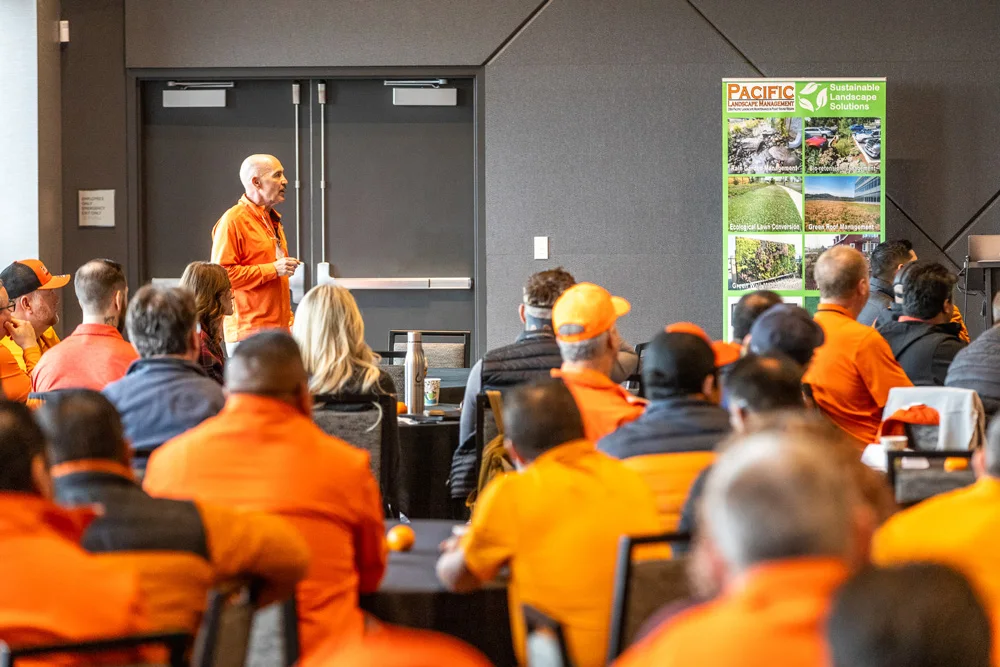 A man in an orange shirt speaks to a group of people, mostly wearing orange, in a conference room with a 
