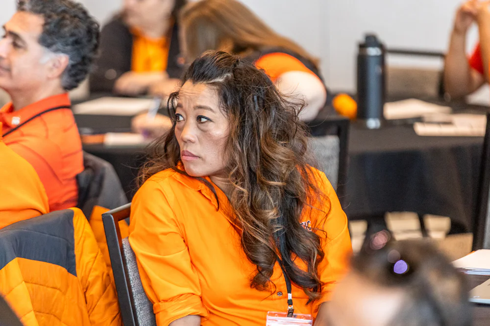 A woman in an orange shirt sits at a conference table, looking to the side attentively. Other attendees in similar shirts are visible in the background, also engaged in the event.