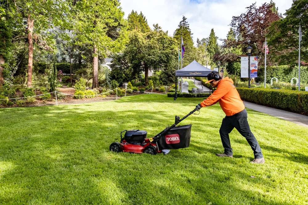 A person wearing an orange jacket and ear protection mows a bright green lawn with a Toro lawnmower in a park surrounded by trees and hedges, with a canopy and banners in the background.