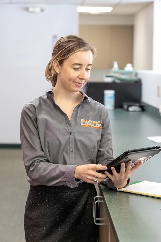 Woman in a gray 'Pacific Landscape Maintenance' shirt standing in an office, leaning on a counter and looking at a tablet. A notepad lies on the counter beside her.