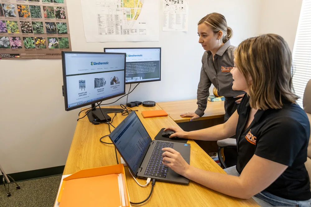 Two women working together at a desk, one sitting and using a laptop with dual monitors, while the other stands looking at the screens. Office setting with wall maps and plant posters.