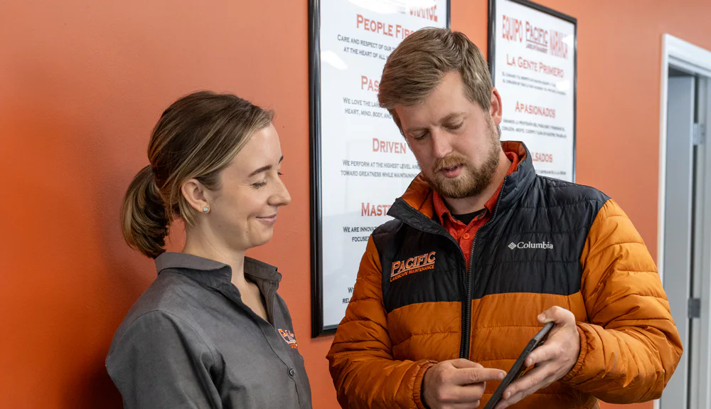 Two employees in work uniforms discuss something while looking at a tablet. They are standing in an office with orange walls and motivational posters behind them.