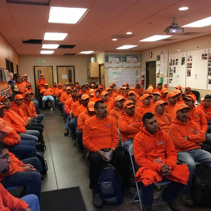 A room filled with workers in matching orange hoodies and caps sitting on chairs and attentively facing forward, suggesting they are in a meeting or training session.