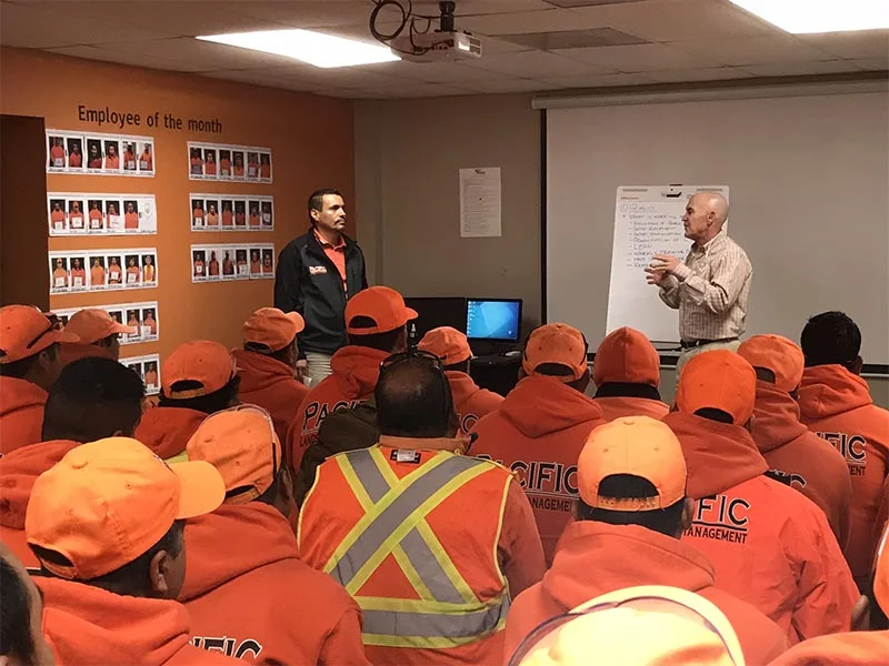 A group of workers in orange uniforms and hats listen to a presentation in a conference room. Two speakers stand at the front, with one writing on a flip chart and an 