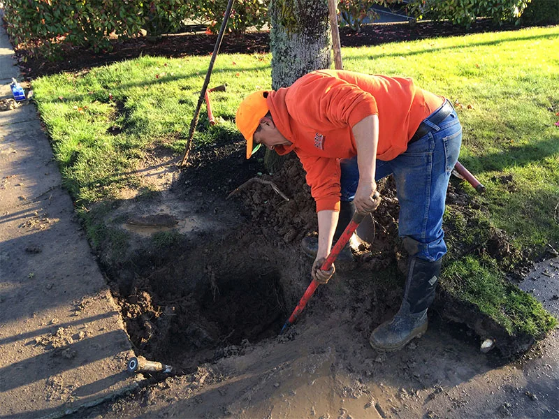 A person wearing an orange hoodie and baseball cap uses a shovel to dig a hole next to a sidewalk. The dirt around the hole is muddy, and grass surrounds the area.