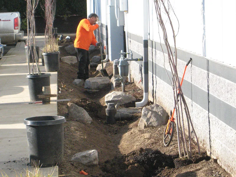 A worker in an orange shirt digs a trench next to a building, surrounded by black potted plants, rocks, and gardening tools. A pickup truck is parked on the left.