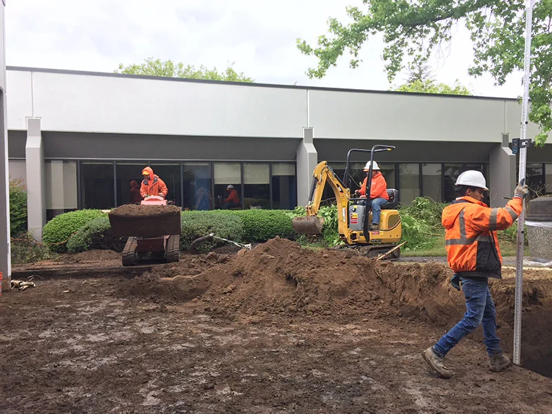 Construction workers in orange jackets and hard hats operate machinery and dig soil outside a building with large windows. One worker uses a mini-excavator while another uses a measuring rod.