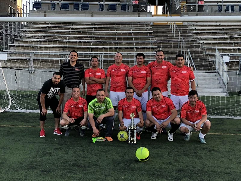 A soccer team in red and white uniforms poses on a field. A trophy and soccer balls are placed in front of them. The background shows bleachers and a goalpost.