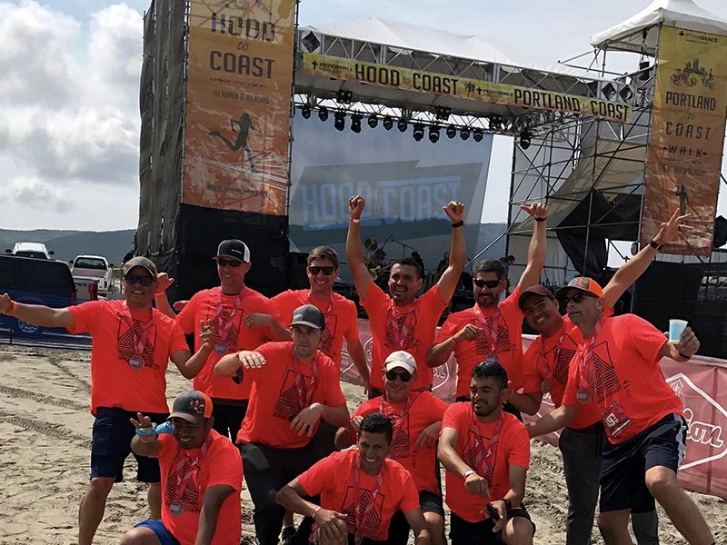 A group of people wearing bright orange shirts celebrate together in front of a Hood-to-Coast event stage on a beach, showing off medals. The stage has banners reading “Hood to Coast” and “Portland to Coast.”