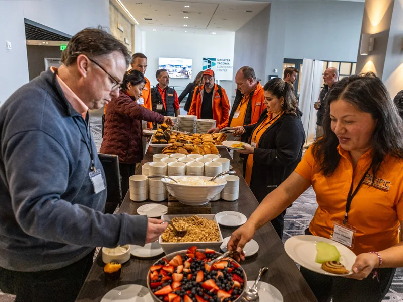 People are gathered around a buffet table with food, including pastries, fruit, and yogurt. Some individuals wear orange shirts and jackets, and the buffet is set in a conference center.