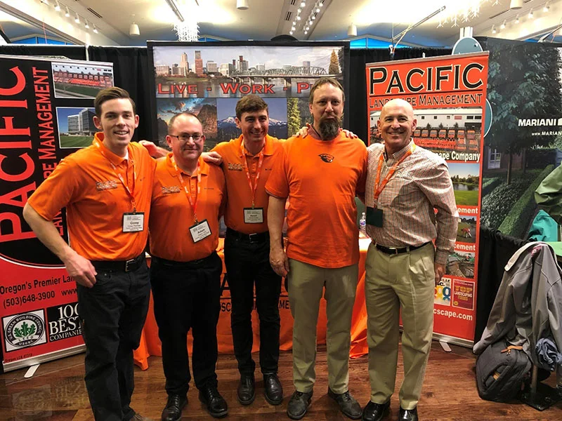 Five men standing closely together in front of a booth featuring banners for Pacific Landscape Management. Four men are wearing orange shirts, and one is wearing a checkered shirt.