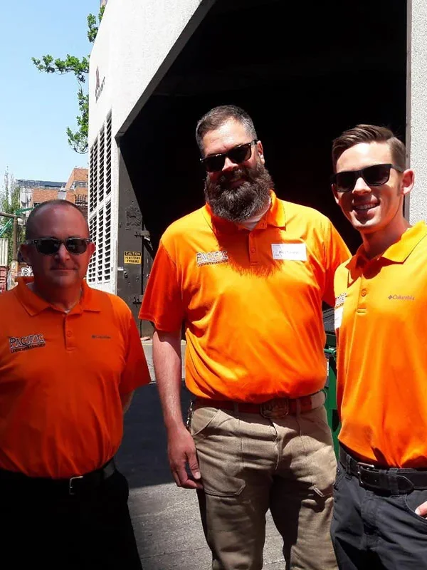 Four people standing outside in bright sunlight wearing sunglasses and orange shirts, smiling at the camera. Trees and a building are visible in the background.