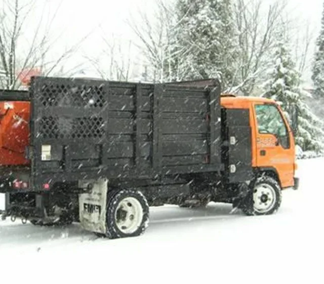 An orange and black utility truck is parked on a snow-covered road with snow falling. Trees covered in snow are visible in the background.