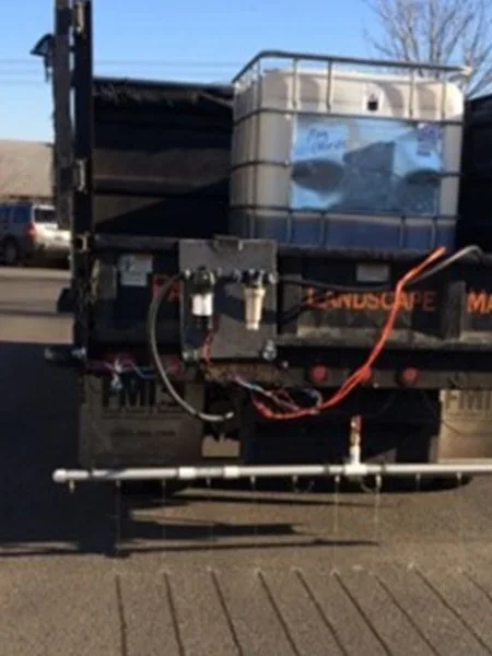 A truck with an open flatbed carrying a large white container and various equipment. The bed has black side panels and the words 