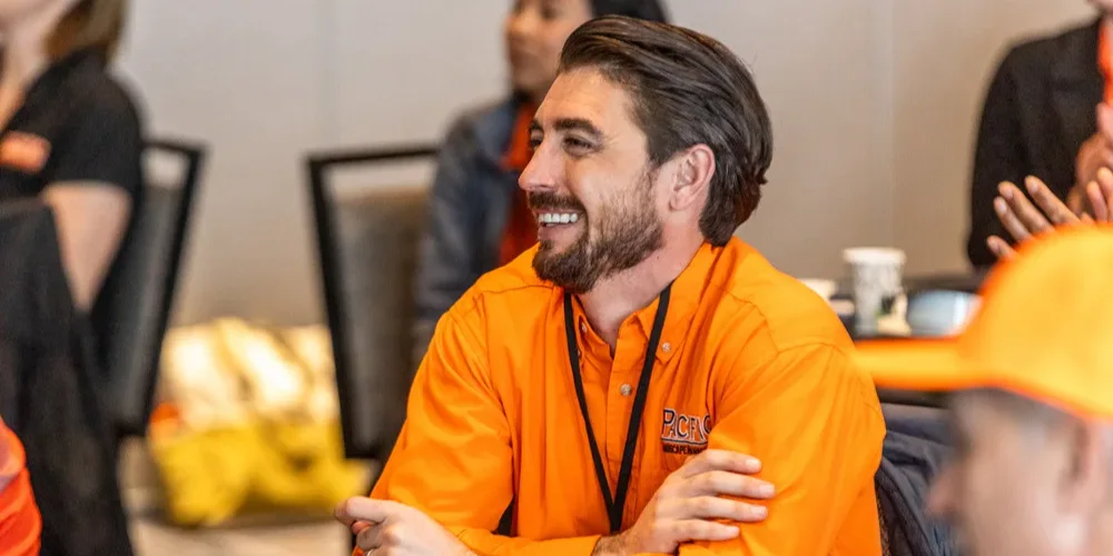 Man in an orange shirt with crossed arms, smiling at a conference table with others. Cup, phone, and plate with food nearby. Office setting in background.