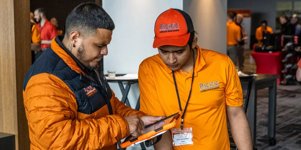 Two men wearing orange Pacific Maintenance uniforms discuss something while looking at a tablet. One man wears a jacket, the other a cap and lanyard with a name tag.