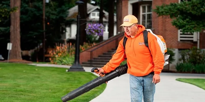 A person in an orange hoodie and hat uses a backpack leaf blower on a grassy area near a walkway, with trees and buildings in the background.