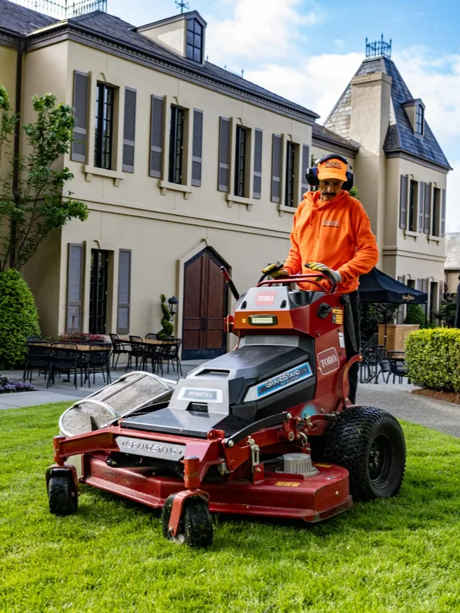 Worker in an orange uniform operates a red Toro Grandstand lawn mower on a manicured lawn in front of a large beige building with multiple windows and outdoor seating.